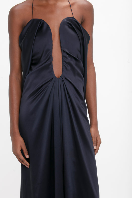 A woman wearing an elegant navy blue evening gown with a plunging neckline and gathered waist detail, focusing on the upper half of the dress and her torso. It is the Victoria Beckham Frame Detail Cut-Out Cami Dress in Navy.