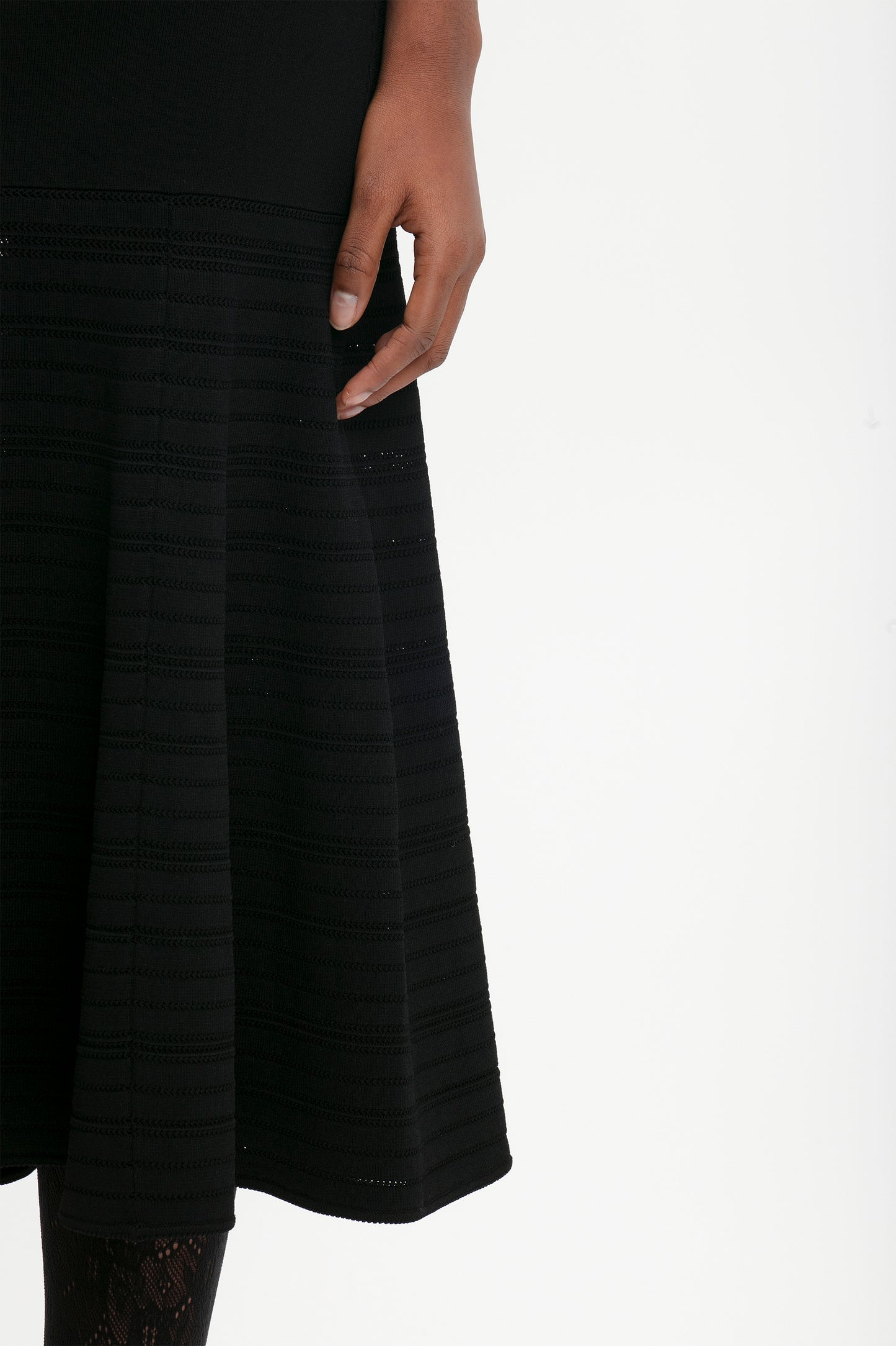 A close-up view of a person’s hand resting on a Fit And Flare Midi Skirt In Black by Victoria Beckham with horizontal lines, against a plain white background. The mid-weight stretch knit clothing features contrasting stitching.