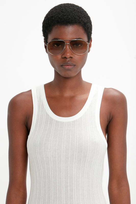 A young woman wearing Victoria Beckham V Metal Pilot sunglasses in Silver-Brown and a white tank top stares directly at the camera against a white background.