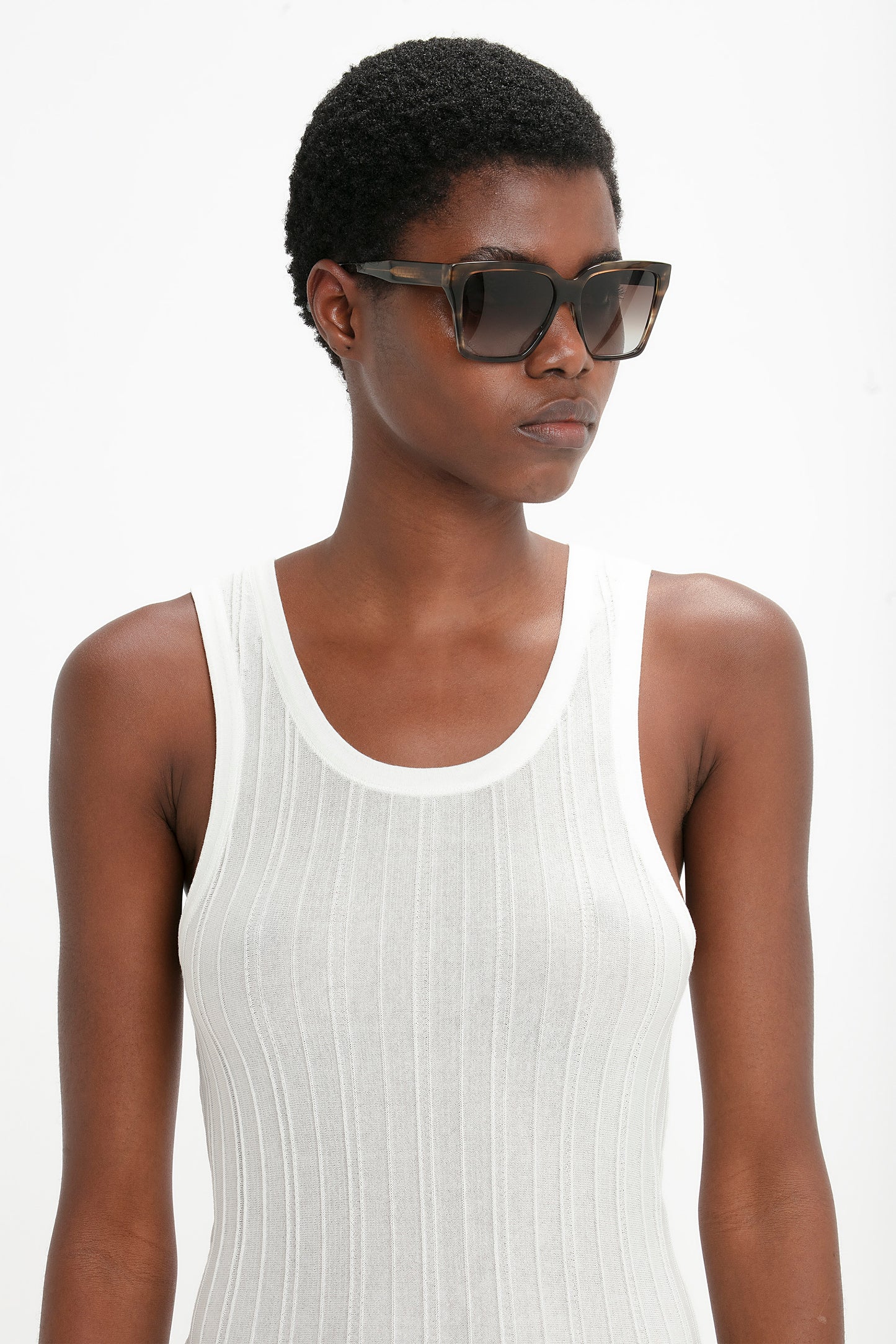 Person with short hair wearing Victoria Beckham Soft Square Frame Sunglasses In Striped Khaki Havana and a white sleeveless ribbed top, standing against a plain background.