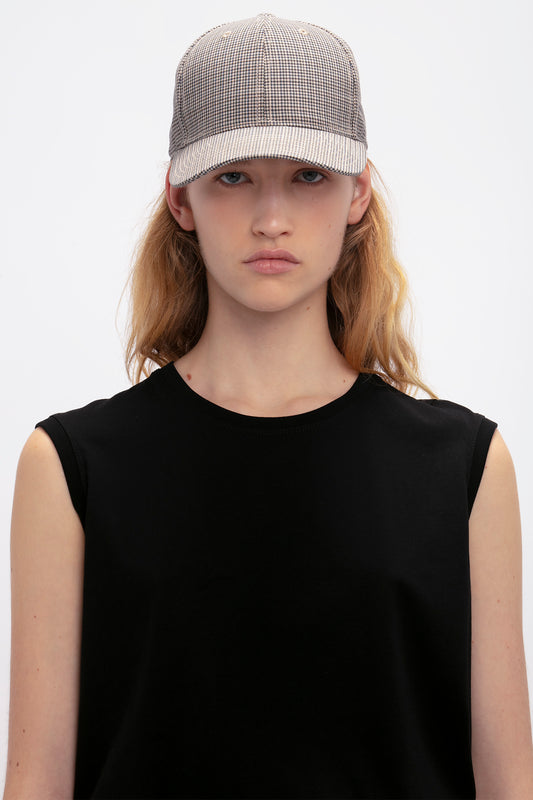 A person with long blonde hair wearing a black top and a wool Victoria Beckham Logo Cap In Dogtooth Check patterned cap, staring directly at the camera.