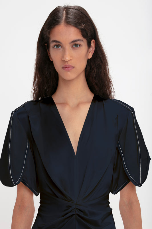 Portrait of a woman with long dark hair, wearing the Victoria Beckham Exclusive Gathered V-Neck Midi Dress in Navy, with puffed sleeves and waist-defining pleat detail.