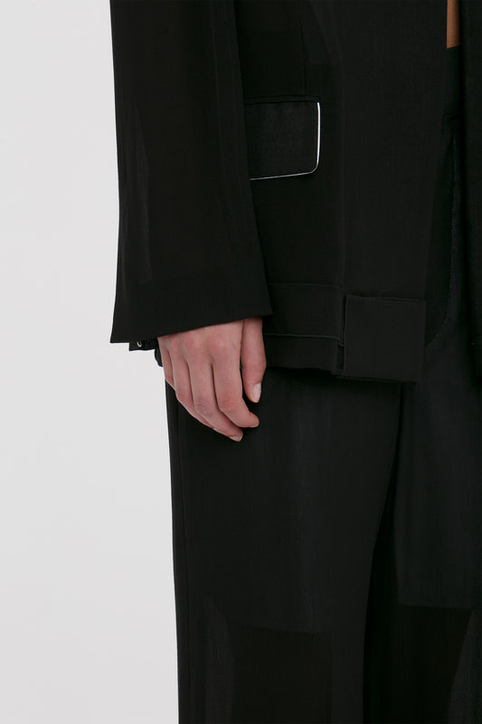 Close-up of a person dressed in a black suit with a focus on their hand hanging beside a Fold Detail Tailored Jacket In Black by Victoria Beckham with a unique waistband detail.
