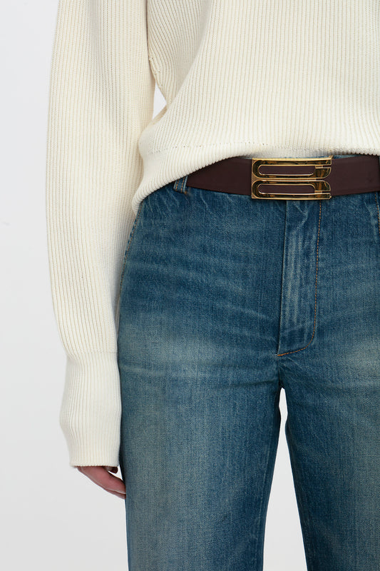 Close-up of a person wearing Victoria Beckham blue Cropped Kick Jean in Indigrey Wash and a cream sweater tucked into a brown belt with a gold buckle, focusing on the midsection.