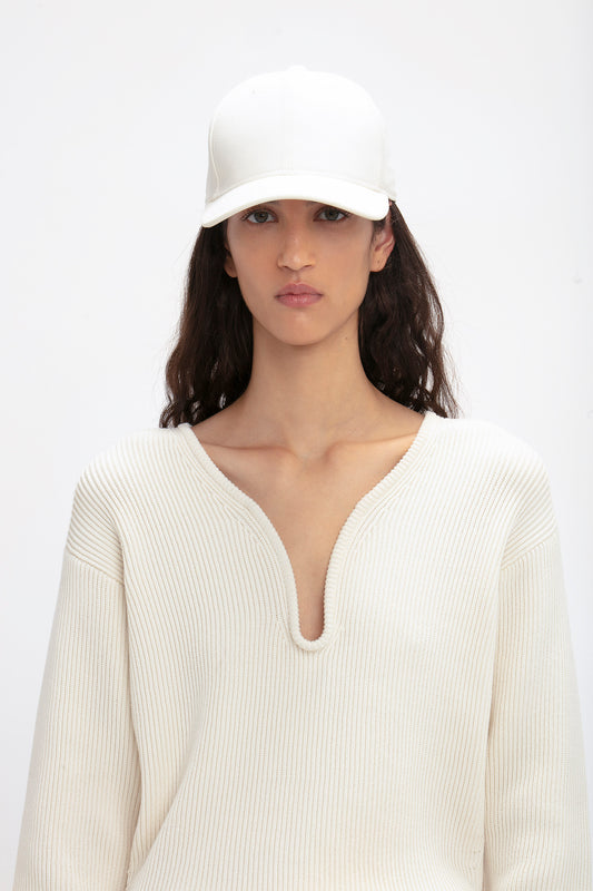 A woman in a white wool crepe sweater and matching Victoria Beckham Logo Cap In Antique White, looking directly at the camera with a neutral expression.