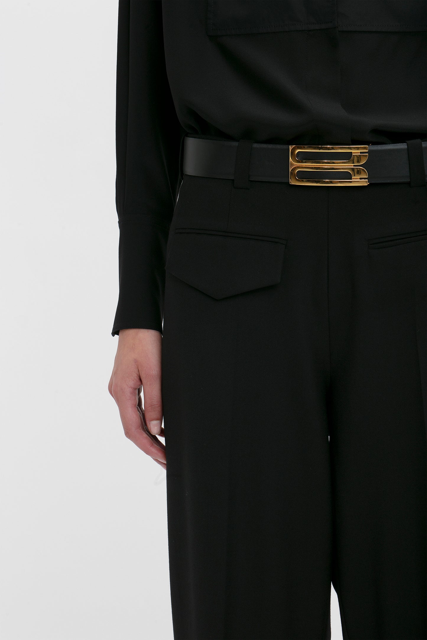 Close-up of a modern woman suiting, with Victoria Beckham's Reverse Front Trouser In Black featuring a gold buckle belt, focusing on the waist area.