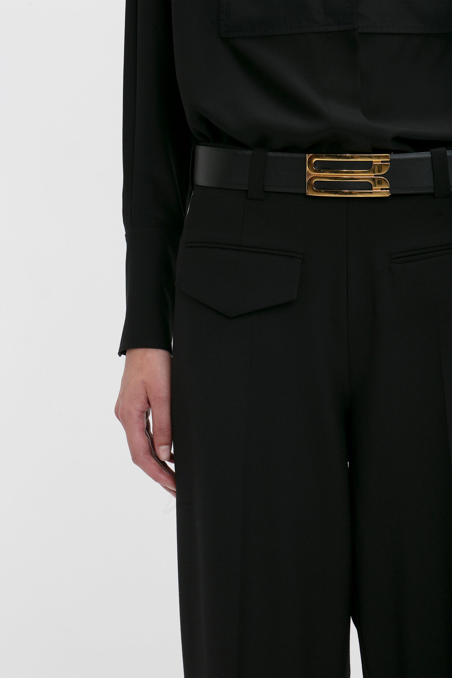 Close-up of a modern woman suiting, with Victoria Beckham's Reverse Front Trouser In Black featuring a gold buckle belt, focusing on the waist area.