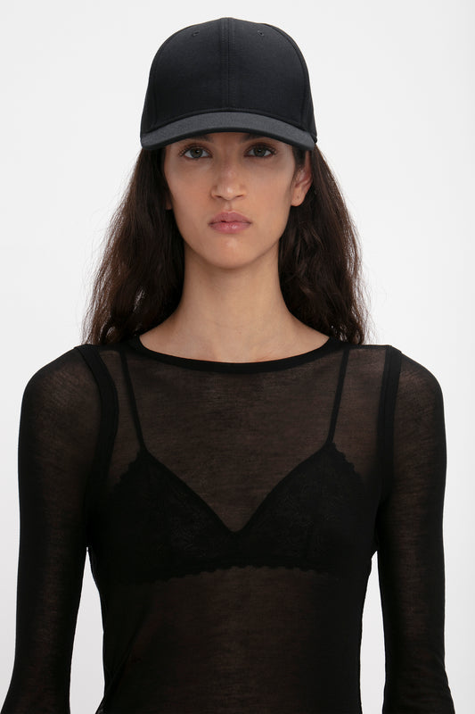 A woman wearing a black mesh top with a lace bra underneath and an Exclusive Logo Cap In Black by Victoria Beckham, posing against a white background.