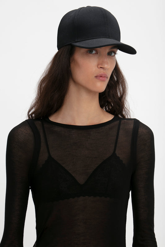 Woman wearing a black Exclusive Logo Cap In Black by Victoria Beckham and sheer top with lace detailing, looking to the side against a white background.