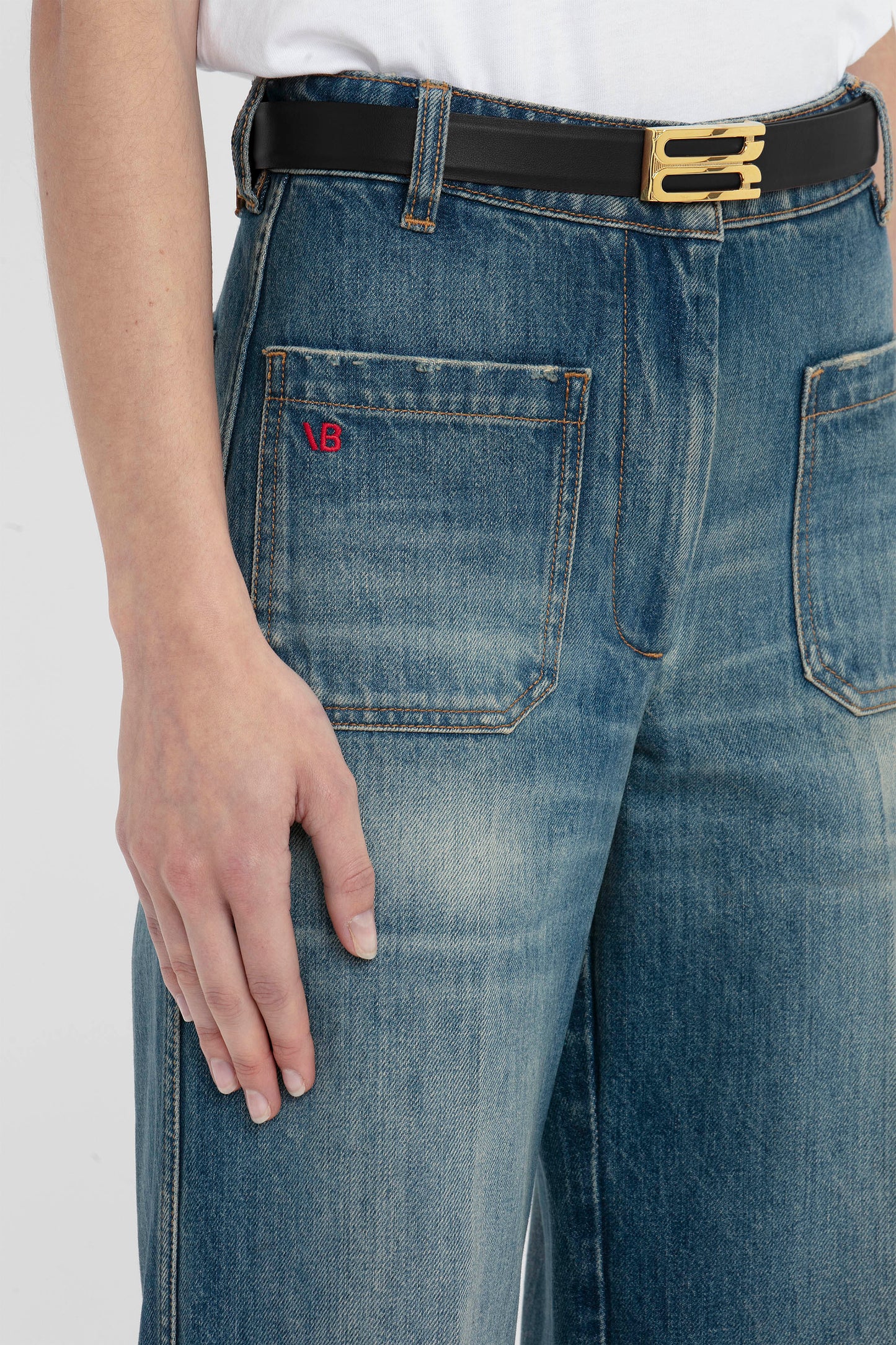 Close-up of a person wearing Victoria Beckham Alina Jean Indigrey Wash blue jeans with a black belt and a red logo on the back pocket, hand resting casually by the side.