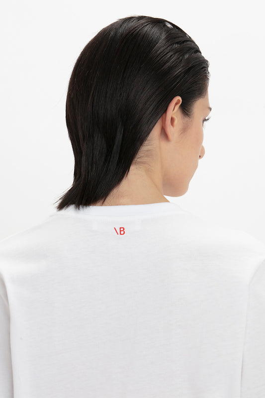 Rear view of a person with dark hair wearing Victoria Beckham's 'David's Wife' Slogan T-Shirt In White made of organic cotton, featuring a small red logo on the collar.