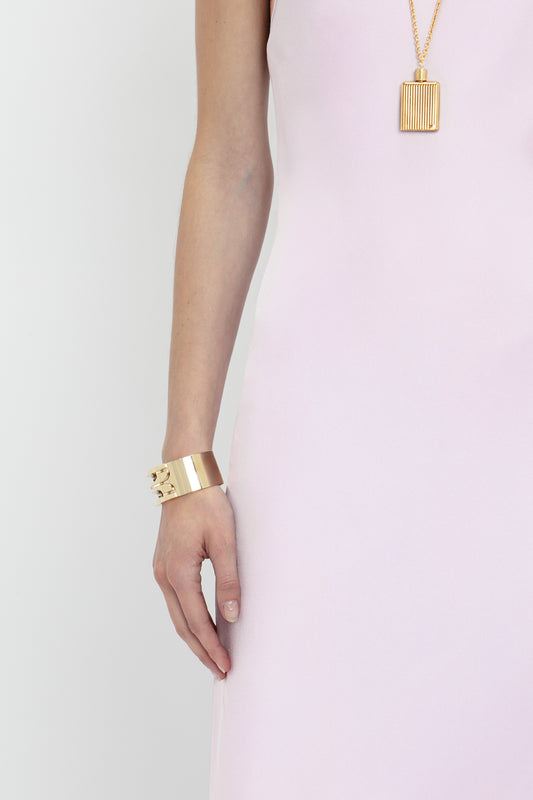 A woman in a Victoria Beckham Low Back Cami Floor-Length Dress In Rosa with a deep V neckline, wearing a gold bracelet and a matching necklace, standing against a grey background.