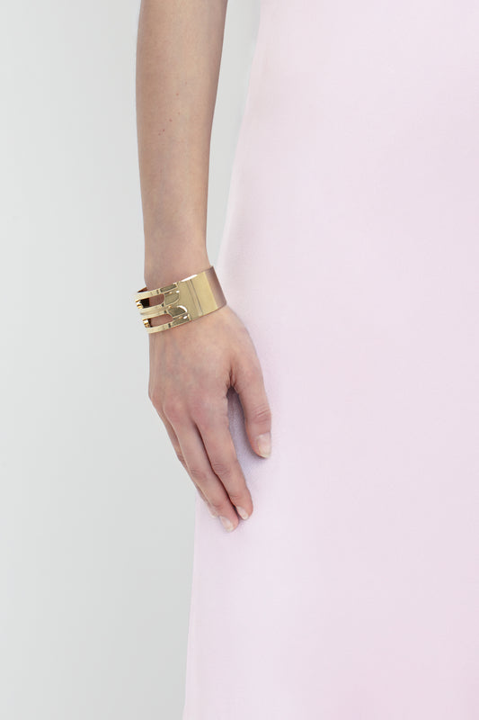 A close-up of a woman's arm by her side, wearing a gold cuff bracelet and a Low Back Cami Floor-Length Dress In Rosa by Victoria Beckham against a gray background.
