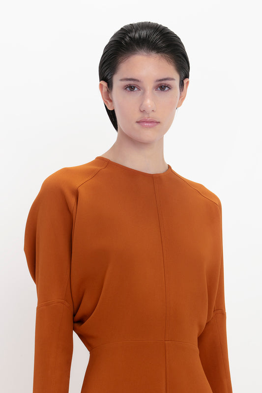 A woman with dark hair styled back, wearing a Victoria Beckham Dolman midi dress in russet, stares directly at the camera against a white background.
