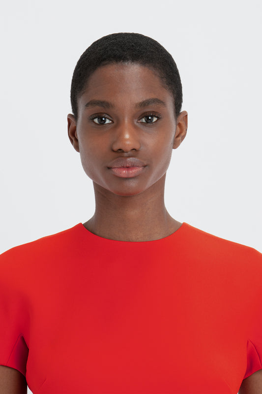 A young woman with short hair, wearing a Victoria Beckham bright red fitted t-shirt dress, standing against a white background, looking directly at the camera.