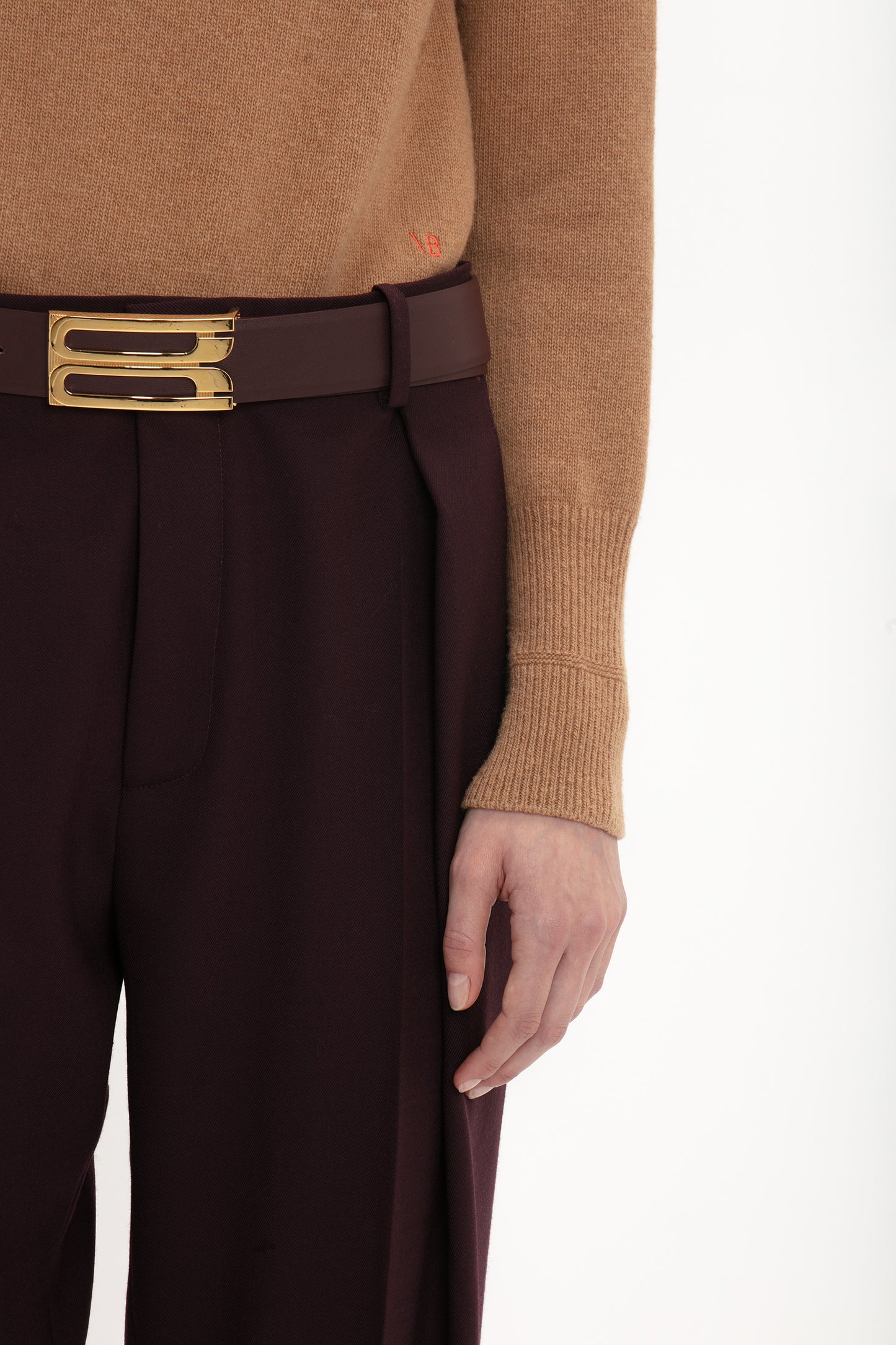 Close-up of a person wearing a lambswool Victoria Beckham polo neck jumper in tobacco and burgundy trousers with a gold buckle belt. Only the torso and hand are visible.