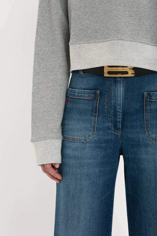 Close-up of a person wearing blue jeans and a Victoria Beckham Cropped Sweatshirt in Grey Marl with their hand resting on their hip, showing an exclusive jumbo frame belt and detailed stitching on the jeans.