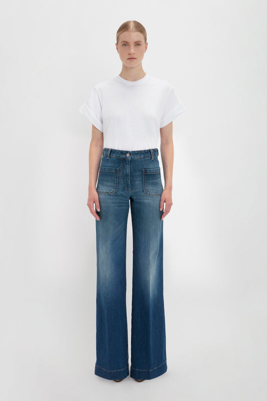 A woman in a Victoria Beckham Asymmetric Relaxed Fit T-Shirt in White and blue jeans standing against a plain background, facing straight ahead with a neutral expression.