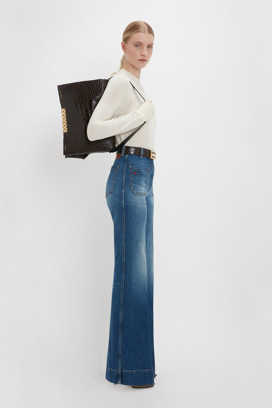 A woman in a Victoria Beckham Merino Crew Jumper In Ivory and blue jeans standing sideways, carrying a large black textured bag.
