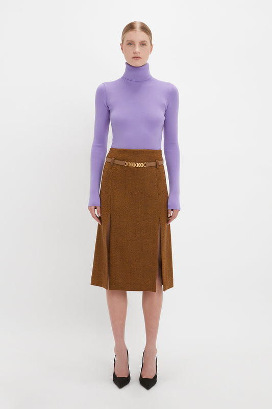 A woman standing against a white background, wearing a lilac knitted polo neck jumper and a brown skirt with a gold chain belt, paired with black pointed heels from Victoria Beckham UK.