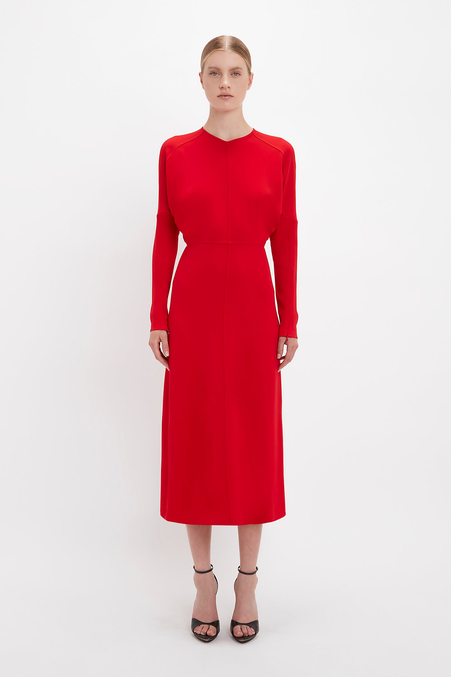 A woman in a simple Victoria Beckham Dolman Midi Red dress and black pointy toe stilettos standing against a white background.
