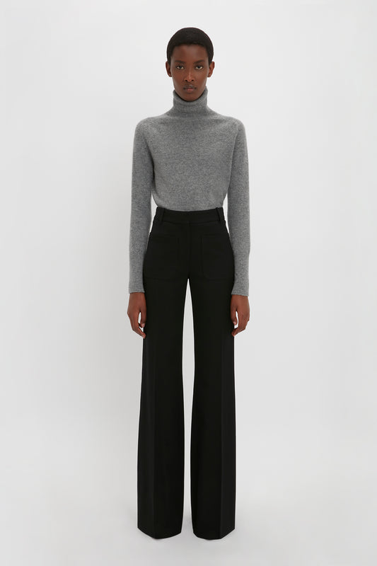 A woman stands against a white background, wearing a grey turtleneck and Victoria Beckham black Alina Tailored Trousers, facing the camera with a neutral expression.