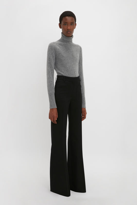 A woman in a grey turtleneck and Victoria Beckham's Alina Tailored Trouser In Black stands against a white background, looking directly at the camera.