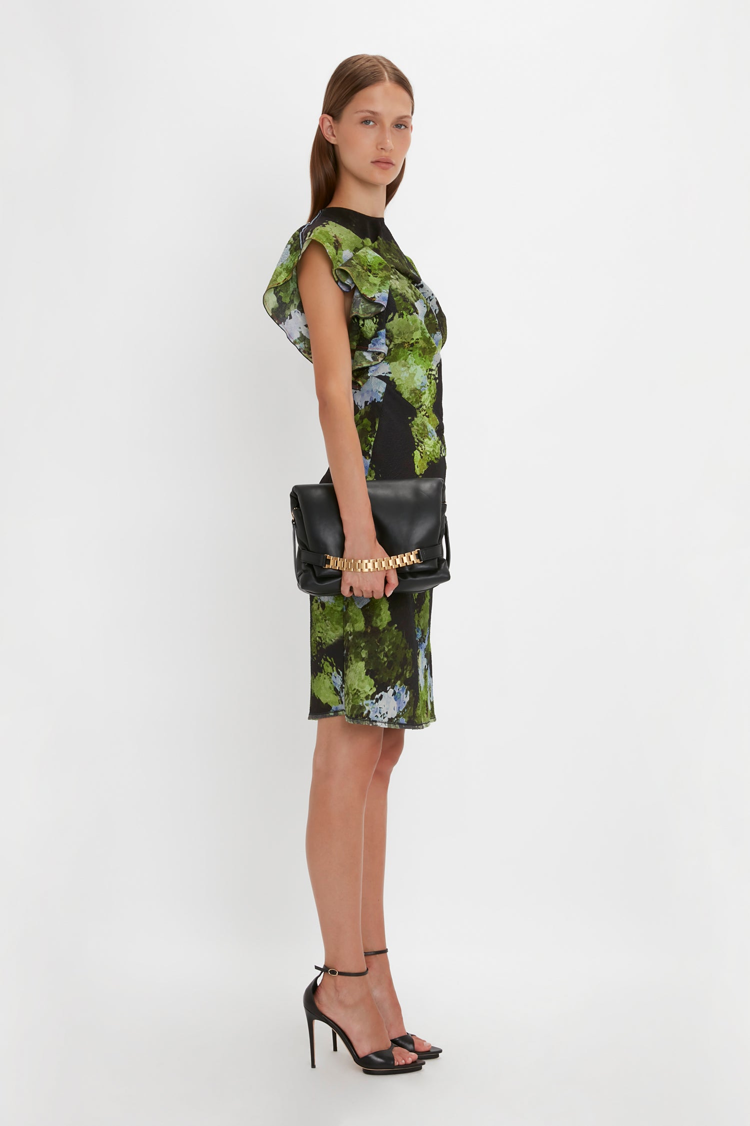 A woman modeling a green and black floral dress with capped sleeves and a Victoria Beckham Puffy Chain Pouch With Strap In Black Leather handbag, standing against a white background.