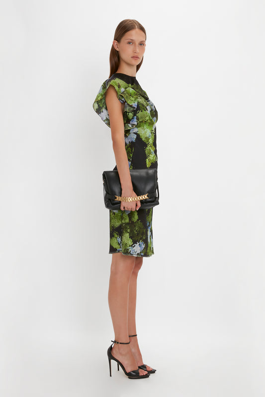 A woman modeling a green and black floral dress with capped sleeves and a Victoria Beckham Puffy Chain Pouch With Strap In Black Leather handbag, standing against a white background.