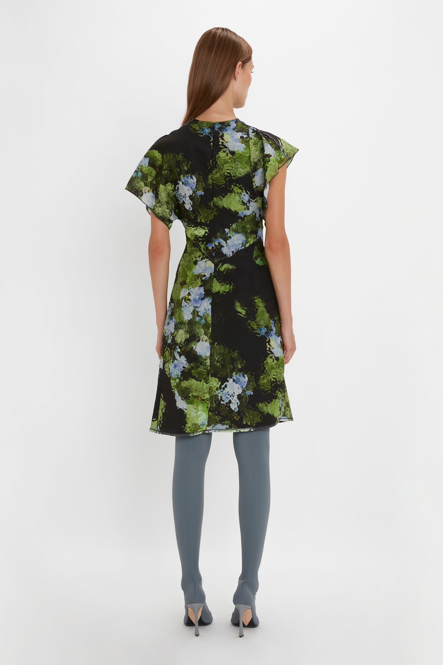 A person is standing with their back to the camera, showcasing a Victoria Beckham Twist Shoulder Dress In Black Frost adorned with green and blue floral patterns. Complementing the waist-defining fit, they pair it with blue stockings and black high-heeled shoes.