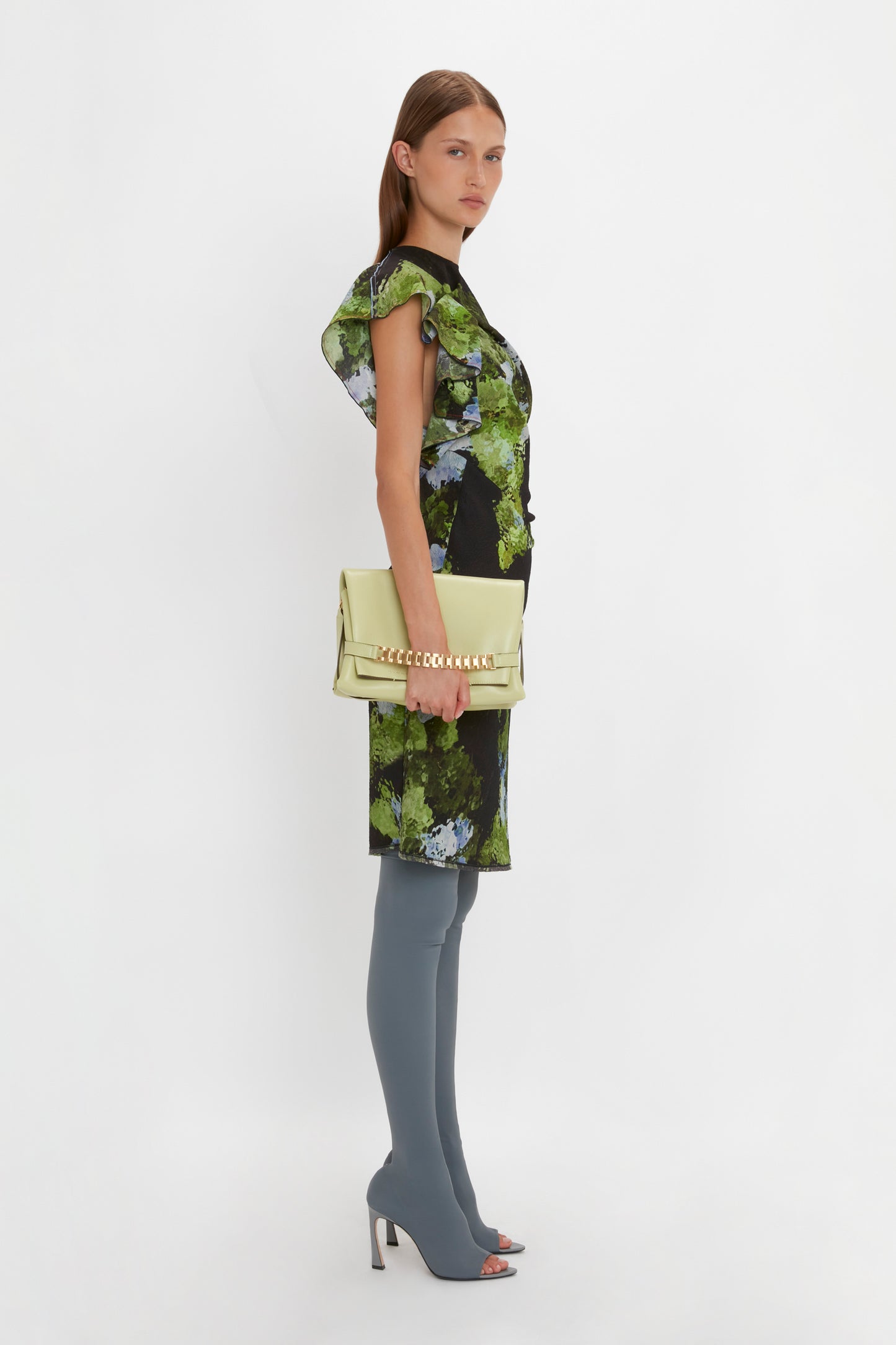A woman stands against a white background wearing a green and black floral Twist Shoulder Dress In Black Frost by Victoria Beckham. She sports knee-high gray boots and holds a light green clutch, accentuating the outfit's waist-defining fit.