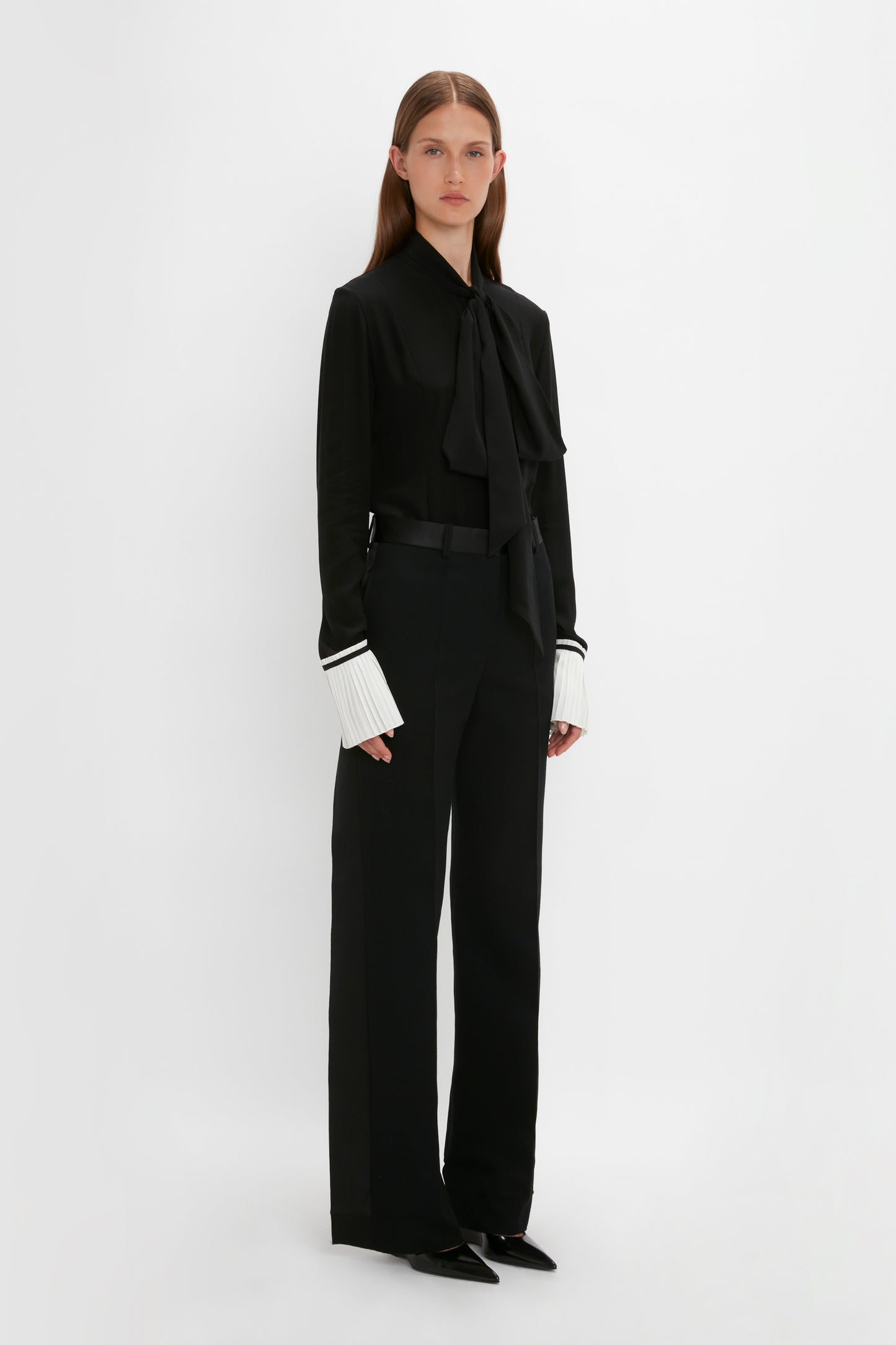 A person stands against a plain white background, wearing the Pleat Cuff Blouse In Black by Victoria Beckham, black trousers, and black pointed shoes.
