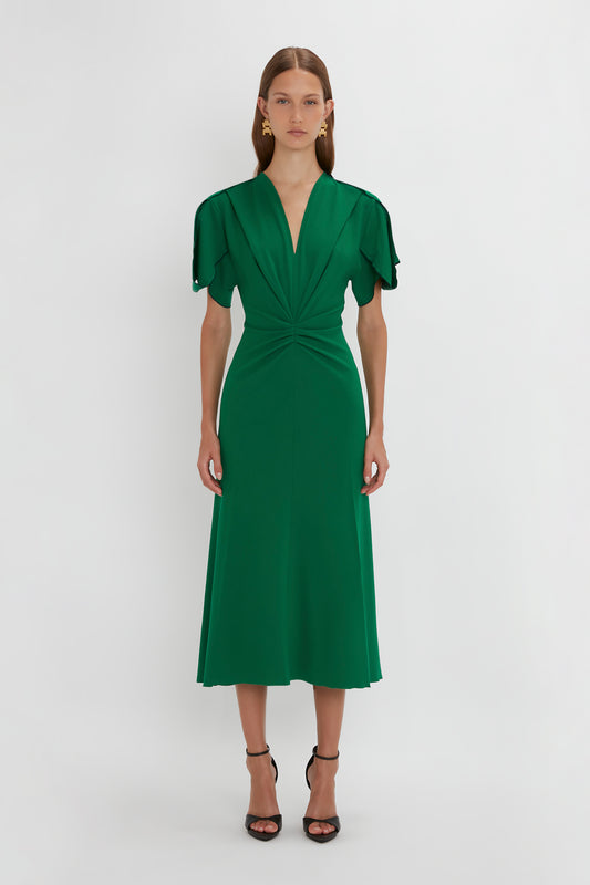 A woman stands against a light background, wearing a Victoria Beckham Gathered V-Neck Midi Dress in Emerald with short sleeves and pointy toe stiletto heels. Her hair is styled back and she has on subtle earrings.