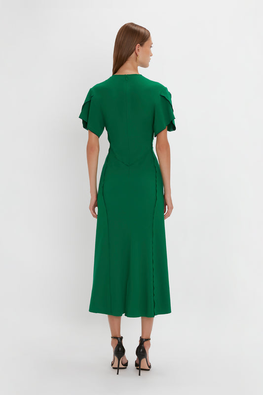 A woman in a Victoria Beckham Gathered V-Neck Midi Dress in Emerald with ruffle sleeves and Pointy Toe Stiletto heels, viewed from behind against a white background.