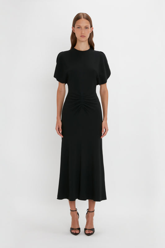 A woman in a black Gathered Waist Midi Dress by Victoria Beckham with short sleeves and a twisted detail at the waist, standing against a white background.