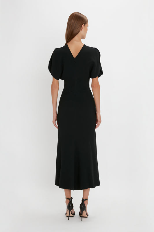 A woman seen from behind, wearing a black, mid-calf length Gathered Waist Midi Dress in Black with short sleeves and standing against a white background.
