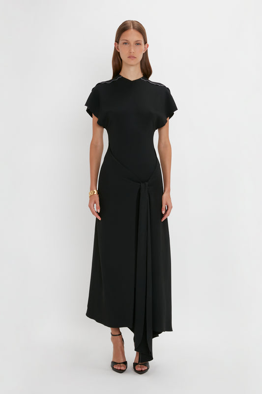 A woman modeling a Victoria Beckham black, elegant dress with short sleeves and a front-tie detail at the waist, standing against a white background.