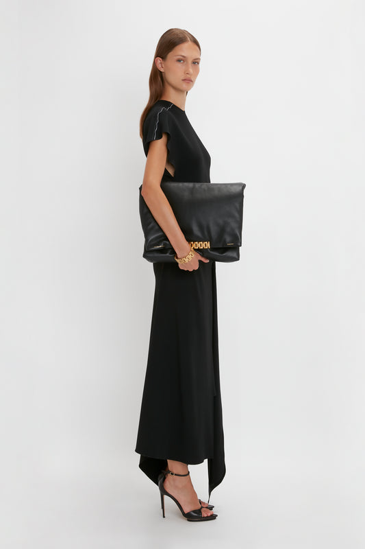 Woman in a Victoria Beckham short sleeve tie detail dress in black holding a large clutch, viewed from the side against a plain white background.