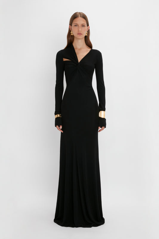 A woman stands against a white backdrop, wearing a long black stretch fabric gown with long sleeves and a twist detail at the chest. She accessorizes with large earrings and gold cuffs by Victoria Beckham.