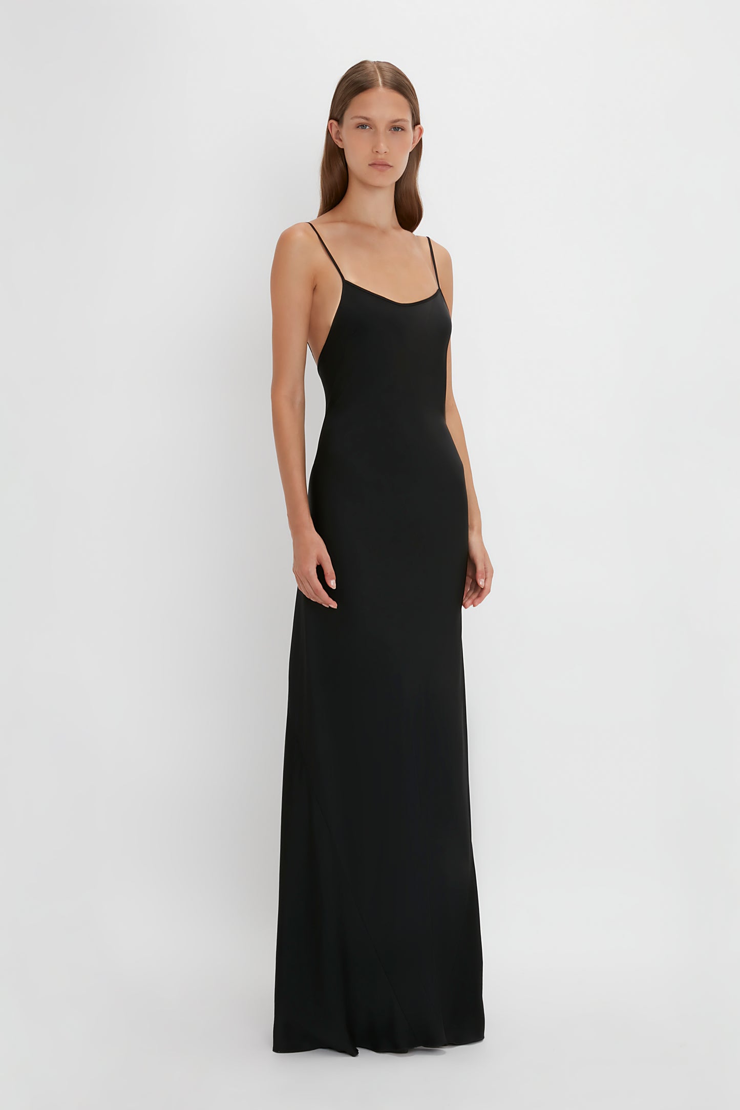 A woman stands against a white background wearing the Victoria Beckham Floor-Length Cami Dress In Black, evoking the elegance of 90s fashion.