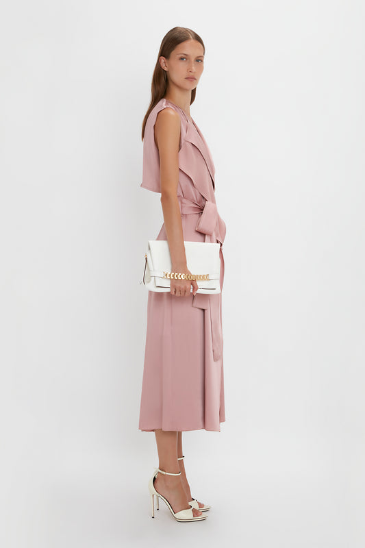 A woman stands in a studio, wearing a pastel pink sleeveless dress with a matching belt and carrying a white Victoria Beckham Nappa leather shoulder bag, complemented by strappy white heels.