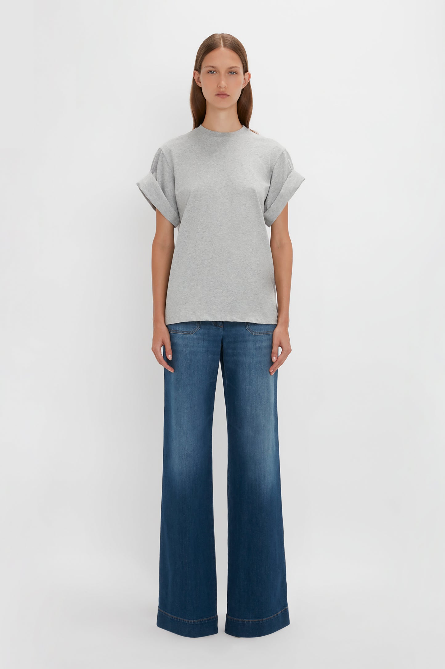 A woman in a Victoria Beckham Asymmetric Relaxed Fit T-shirt in Grey Marl and blue flared jeans standing against a white background.