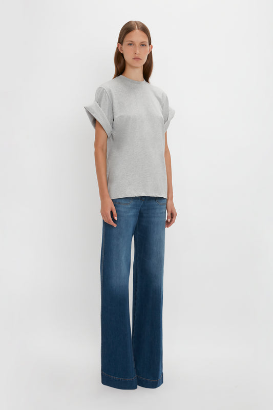 Woman in a Victoria Beckham Asymmetric Relaxed Fit T-Shirt In Grey Marl and blue jeans standing against a white background.