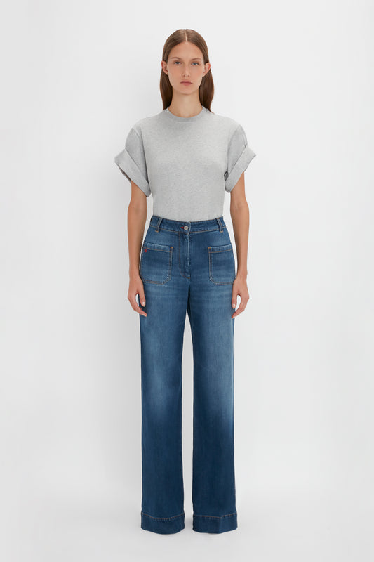 A woman in a studio setting wearing a Victoria Beckham Asymmetric Relaxed Fit T-shirt in Grey Marl tucked into high-waisted blue jeans.