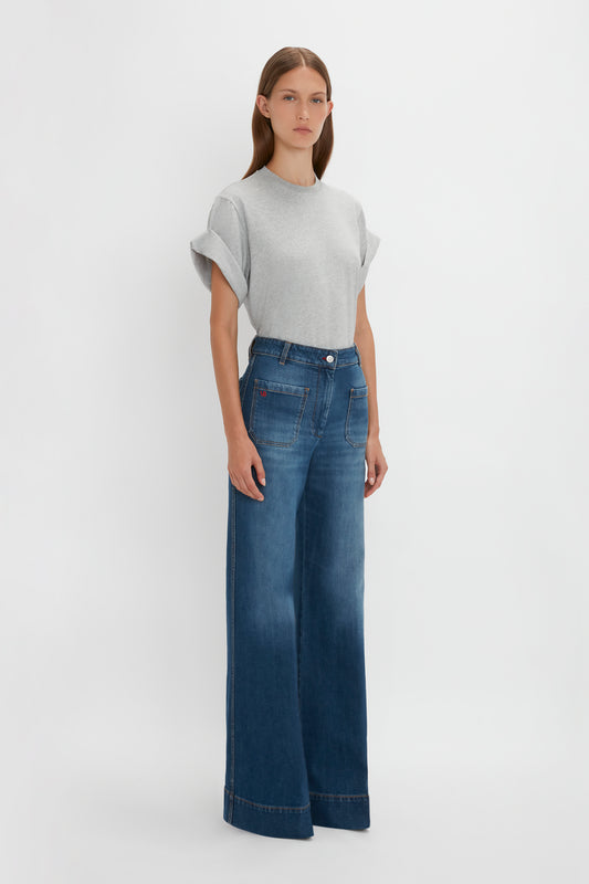 A woman in a Victoria Beckham Asymmetric Relaxed Fit T-Shirt In Grey Marl and blue wide-leg jeans stands against a white background.