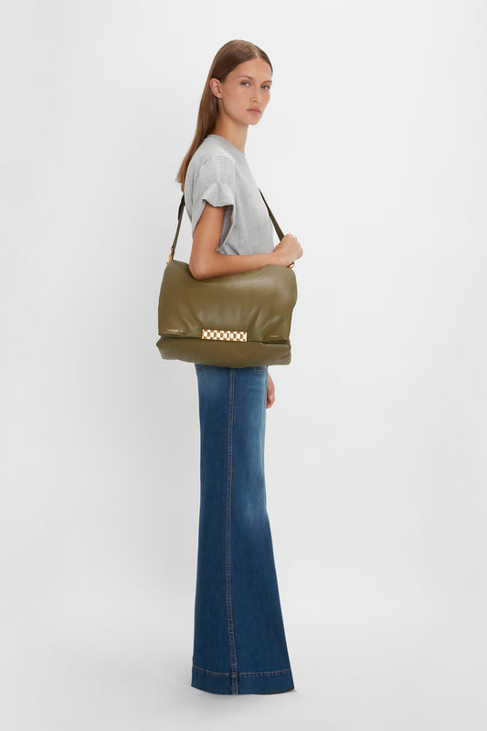 A woman in a gray t-shirt and Victoria Beckham Alina Jean in Dark Vintage Wash carrying a large olive green shoulder bag, standing against a white background.