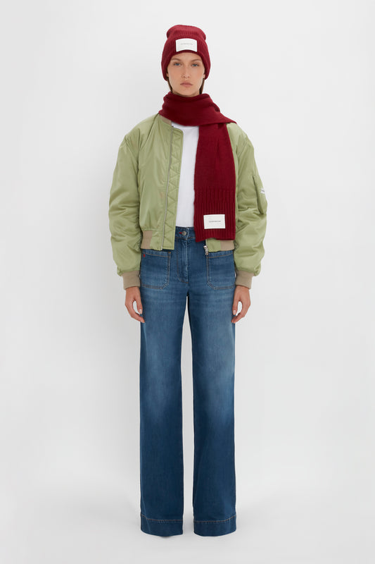 A person in a green bomber jacket, red beanie, and a Victoria Beckham Exclusive Logo Patch Scarf In Burgundy, wearing light blue jeans stands facing forward against a white background.