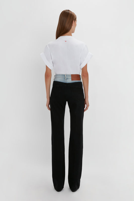 A woman stands facing away, wearing a white t-shirt and black high-waisted, relaxed leg Julia Jeans In Contrast Wash by Victoria Beckham against a plain background.