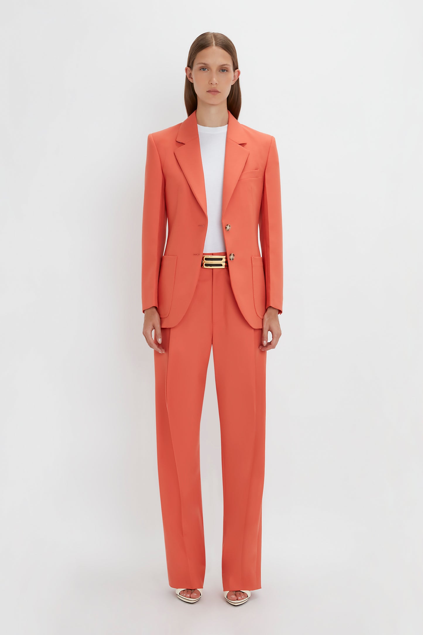 A woman stands against a plain background wearing a Victoria Beckham orange single-breasted jacket in papaya with matching single pleat trousers, complemented by silver sandals.