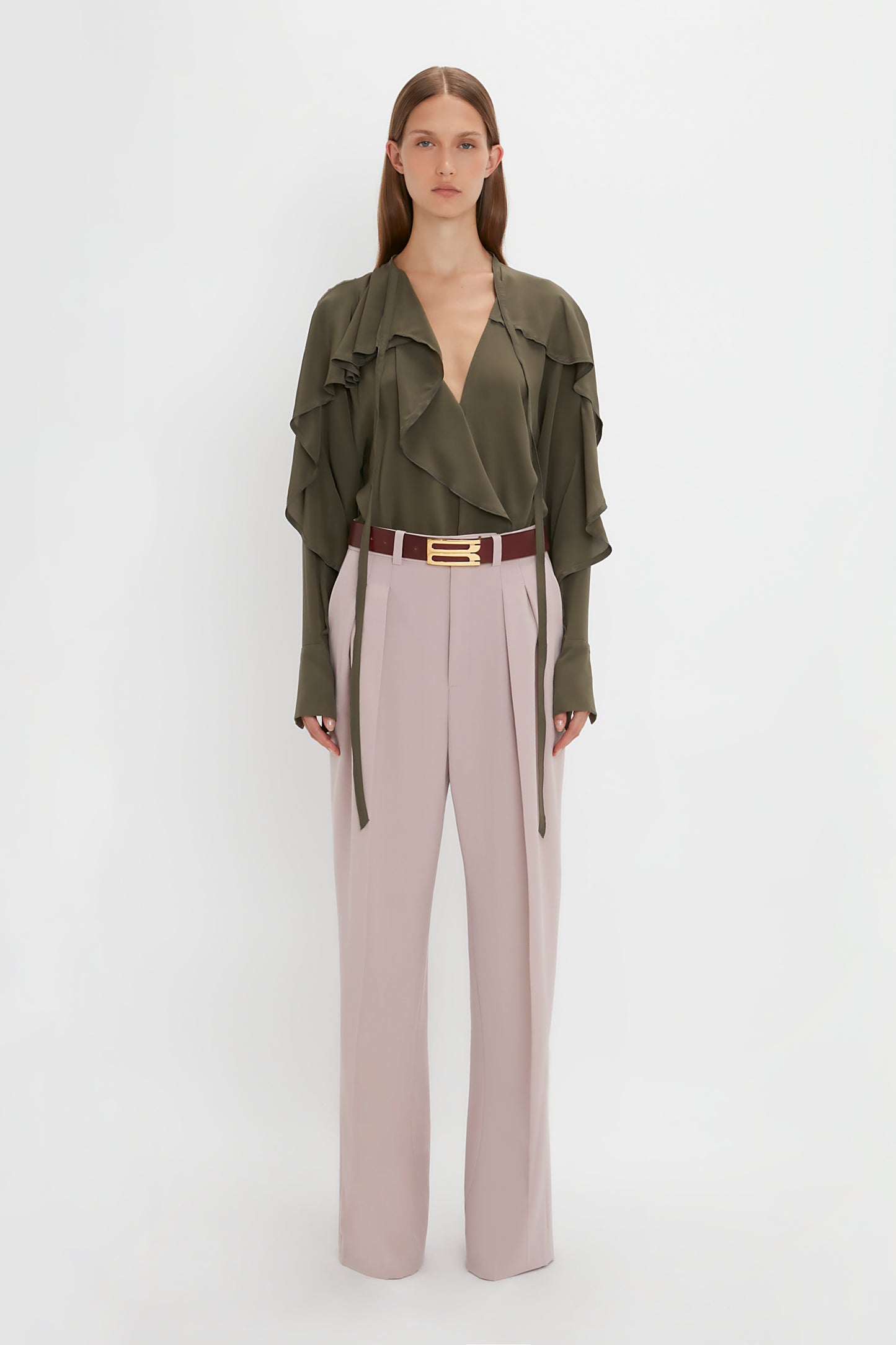 A person stands facing the camera wearing a green **Tie Detail Ruffle Blouse in Oregano by Victoria Beckham** and light pink Double Pleat Trousers with a brown belt. The background is plain white.